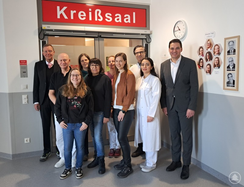 Landrat Sebastian Gruber und Kliniken Geschäftsführer Marcus Plaschke freuen sich gemeinsam mit dem Geburtshilfeteam aus Ärzten, Belegärzten und Hebammen über die erneute Förderung. Landrat Sebastian Gruber und Klinikgeschäftsführer Marcus Plaschke stehen gemeinsam mit Ärzten, Hebammen und Pflegepersonal vor dem Eingang zum Kreißsaal im Krankenhaus Freyung.