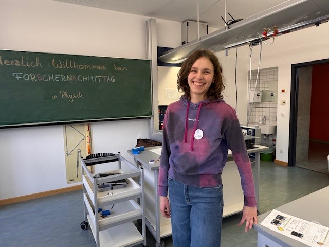 Tutorin begrüßt die Teilnehmerinnen und Teilnehmer zum Forschernachmittag im Physikraum. Eine Schülerin steht lächelnd im Physikraum vor einer Tafel mit der Aufschrift Willkommen beim Forschernachmittag in Physik.