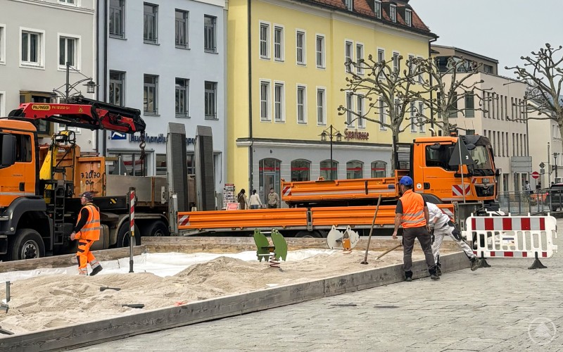Vorbereitungen für die sommerliche Sandinsel laufen auf Hochtouren Bauarbeiter in Warnkleidung bereiten auf dem Stadtplatz eine große Sandfläche mit Spielgeräten vor, im Hintergrund stehen Lkw und Gebäude der Innenstadt.