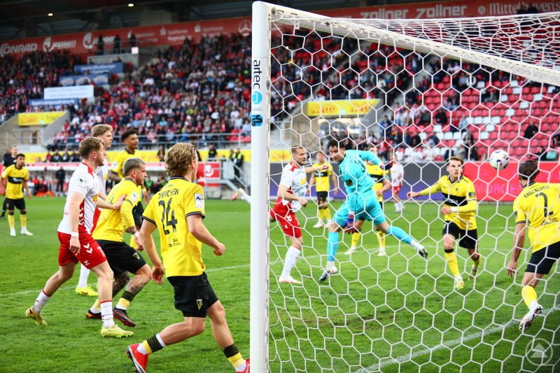 Der Ball zappelt im Netz: Aachen trifft im Strafraumgewühl vor zahlreichen Zuschauern zur Wende in Regensburg. Szene im Strafraum: Mehrere Spieler stehen dicht vor dem Tor, der Ball fliegt Richtung Netz. Der Torwart reagiert, während Zuschauer im Hintergrund das Geschehen verfolgen.