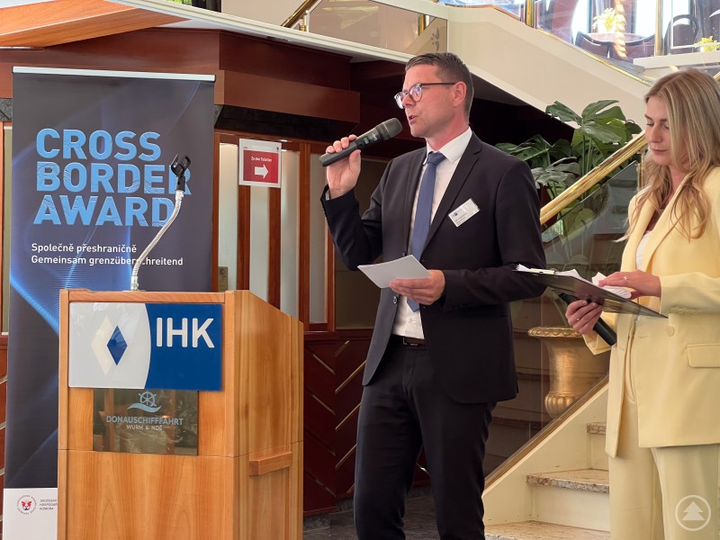 Begrüßung der Gäste durch IHK-Vizepräsident Jürgen Greipl beim Cross Border Award 2024. Ein Mann im Anzug spricht an einem Rednerpult mit IHK-Logo in ein Mikrofon. Neben ihm steht eine Frau im hellgelben Hosenanzug und hält ein Mikrofon und Unterlagen. Hinter dem Rednerpult befindet sich ein Banner mit der Aufschrift „CROSS BORDER AWARD“. Bildunterschriften: