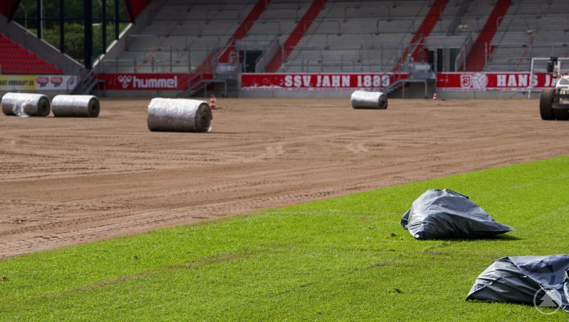 Mehrere Rollen Rollrasen liegen auf vorbereiteter Erde in einem Stadion, ein Teil des Rasens ist bereits verlegt.