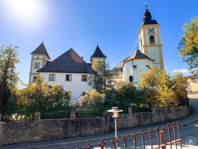 Auch kulturelle Highlights wie das Kloster Rinchnach werden im WALDgeist vorgestellt. Barocke Klosteranlage mit markantem Turm und sonniger Fassade, umgeben von Bäumen.