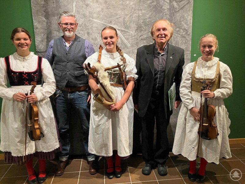Bei der Vernissage: Christian Binder, Leiter des Waldgeschichtlichen Museums (l.), und Dr. Vladimir Horpeniak, ehemaliger Leiter des Muzeum Šumavy, mit dem „Lustigen Dudelsacktrio“. Drei junge Musikerinnen in traditioneller Tracht mit Geige und Dudelsack stehen gemeinsam mit zwei Männern in Anzug bzw. Weste vor einer Ausstellungswand. Alle lächeln in die Kamera.
