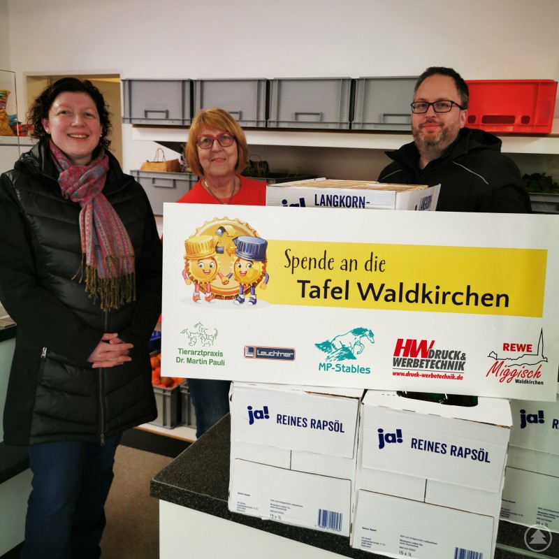Spendenübergabe an die Tafel Waldkirchen (von links) Sigrid Donaubauer (MP Stables), Christine Lang (Tafel Waldkirchen) und Herbert Weiß (HW Druck & Werbetechnik). Drei Personen stehen in einem Raum mit Kisten und Lebensmitteln. Sie halten ein großes Schild mit der Aufschrift Spende an die Tafel Waldkirchen.
