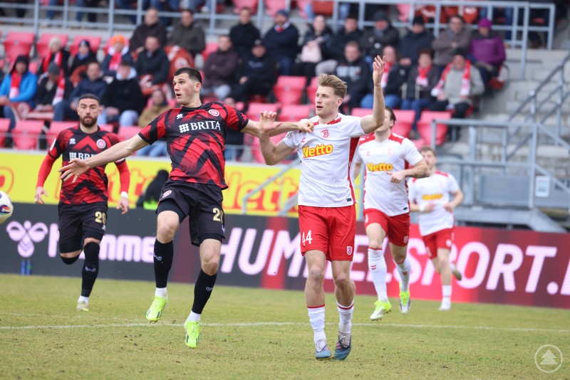 Zwei Spieler springen im Laufduell um den Ball, im Hintergrund verfolgen Zuschauer auf roten Sitzen die Szene.