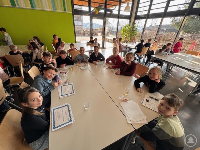 Zum Abschluss erhielten alle Teilnehmerinnen und Teilnehmer ein Einstein Forscherdiplom. Viele Grundschulkinder sitzen gemeinsam an großen Tischen in einem hellen Raum und halten ihre Forscherdiplome in den Händen.