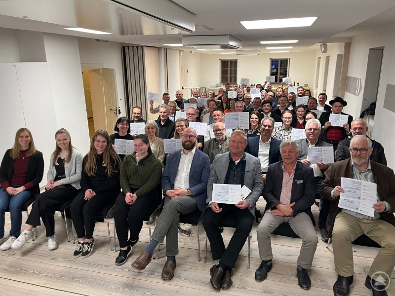 Knapp 60 Vereinsvertreterinnen und -vertreter sowie Bürgermeister freuen sich über die finanzielle Unterstützung für 26 Projekte im Ilzer Land. Eine große Gruppe von Vereinsvertretern, Bürgermeistern und Organisatoren sitzt in einem Veranstaltungsraum und hält Förderurkunden in die Kamera. Die Teilnehmer lächeln und präsentieren stolz ihre Auszeichnungen.