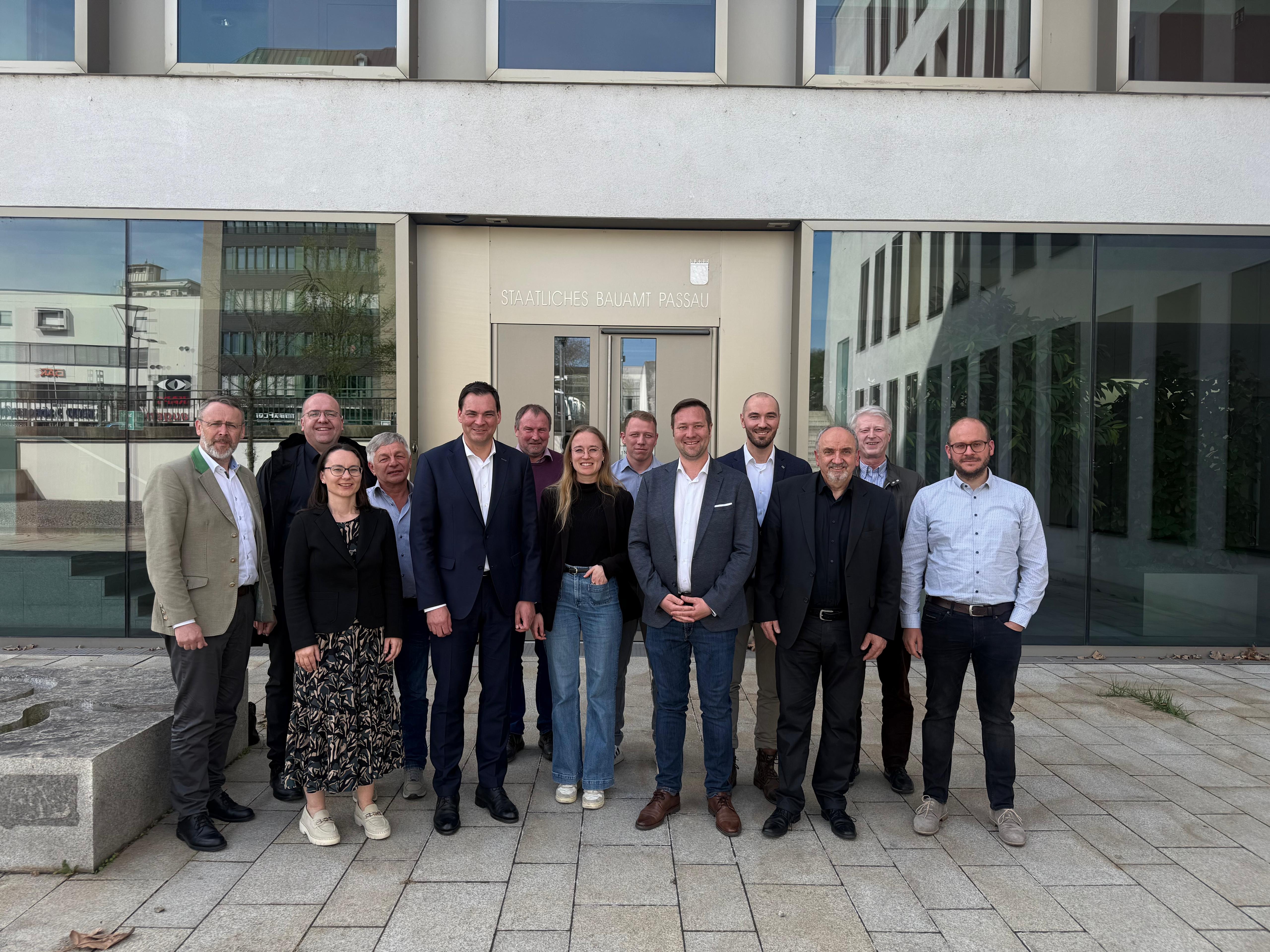 Das Bild zeigt eine Gruppe von 14 Personen, die sich vor dem Gebäude des Staatlichen Bauamts Passau versammelt haben. Die Personen sind unterschiedlich gekleidet, einige formell in Anzügen und andere etwas legerer. Das Gebäude hat eine moderne Glasfassade mit einem klar erkennbaren Schild, das den Namen 