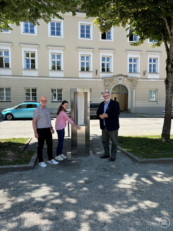 Oberbürgermeister Jürgen Dupper (rechts) nimmt gemeinsam mit Wolfgang Seiderer (links, Referent für Bau- und Umwelt) und Carolin Stadler (Mitte, Umweltschutz Stadt Passau) den neuen Trinkwasserbrunnen am Domplatz in Betrieb. Drei Personen stehen unter Bäumen neben einem modernen Edelstahl-Trinkwasserbrunnen auf einem Platz vor einem historischen Gebäude mit hellbeiger Fassade.