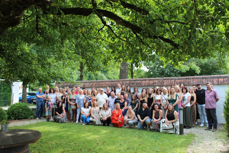 Mitte: Geschäftsführer Herr Christoph Liebl und Herr Heiko Schumann mit stellvertretender GF Verena Liebl; Mitarbeiterinnen und Mitarbeiter der Firma SoNet Eine große Gruppe von Mitarbeiterinnen und Mitarbeitern steht gemeinsam in einem Garten unter einem großen Baum. Im Hintergrund ist eine Mauer und viel Grün zu sehen. Die Personen lächeln und posieren für ein Gruppenfoto.