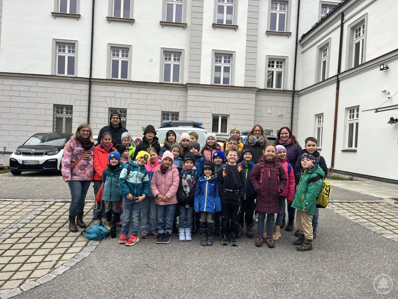 Die Kinderfeuerwehr Bodenmais mit Polizeihauptmeister Maximilian Dengler und den Betreuerinnen Barbara Vogl, Marina Müller und Evi Trauner bei ihrem Besuch in der Polizeiinspektion Regen. Eine Gruppe von Kindern der Kinderfeuerwehr Bodenmais steht gemeinsam mit ihren Betreuerinnen und einem Polizeibeamten im Hof der Polizeiinspektion Regen. Alle tragen wetterfeste Kleidung und lächeln in die Kamera.