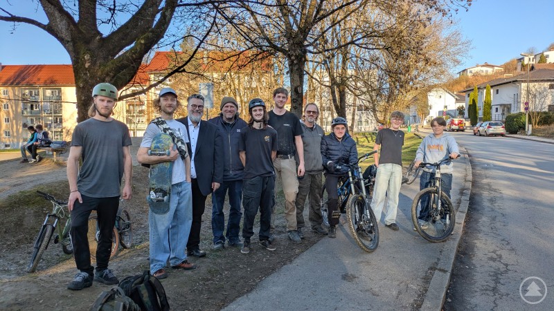 Vor Ort informierten sich Bürgermeister Andreas Rother (3. von links), Andreas Schützenberger von IOU-Ramps (4. von links) sowie Vertreter der örtlichen Dirt-Bike-Community über die geplante Aufwertung des Dirt-Bike-Parks an der Westerburgerstraße. Eine Gruppe junger Mountainbiker steht gemeinsam mit Vertretern der Stadt und Fachleuten an einem Dirt-Bike-Park. Einige halten ihre Fahrräder, andere tragen Helme oder halten ein Skateboard. Im Hintergrund sind Bäume und Wohnhäuser zu sehen.