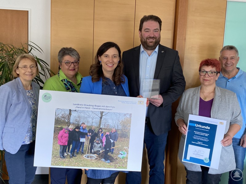 Präsentierten die Auszeichnung Landrat Tobias Beck (3. von rechts): Brigitte Tschimmel (Leiterin Sachgebiet ÖPNV, Mobilität, Kreisentwicklung), Gerlinde Gietl (Leiterin Amt für Jugend und Familie), Julia Stadler (Kommunale Jugendpflegerin), Barbara Herrnberger (Seniorenfachstelle) und Richard Maier (Geschäftsführer Kreisjugendring, von links). Mehrere Vertreterinnen und Vertreter des Landkreises Straubing-Bogen präsentieren gemeinsam eine Auszeichnung und ein prämiertes Foto zum Projekt „Hand in Hand – Generationenbaum“.