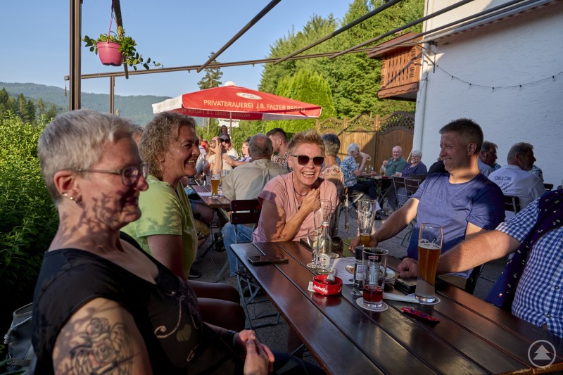 Eine fröhliche Gruppe sitzt im Biergarten an einem Holztisch, lacht und trinkt bei schönem Wetter.