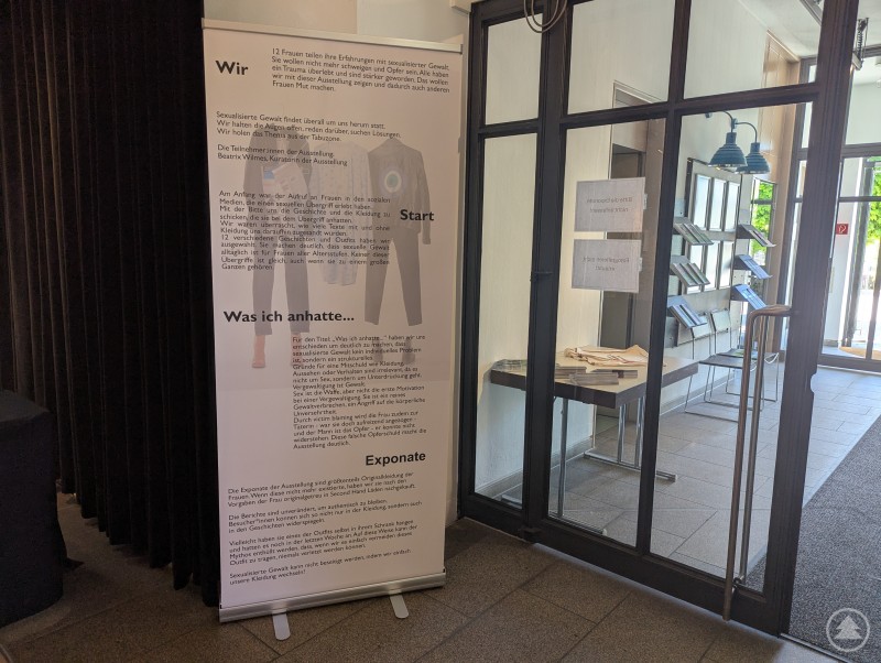 Das Bild zeigt ein Roll-Up-Banner einer Ausstellung, in der 13 Frauen ihre Erfahrungen mit sexualisierter Gewalt teilen und Kleidung zeigen, die sie während des Übergriffs trugen, um mit Vorurteilen aufzuräumen.