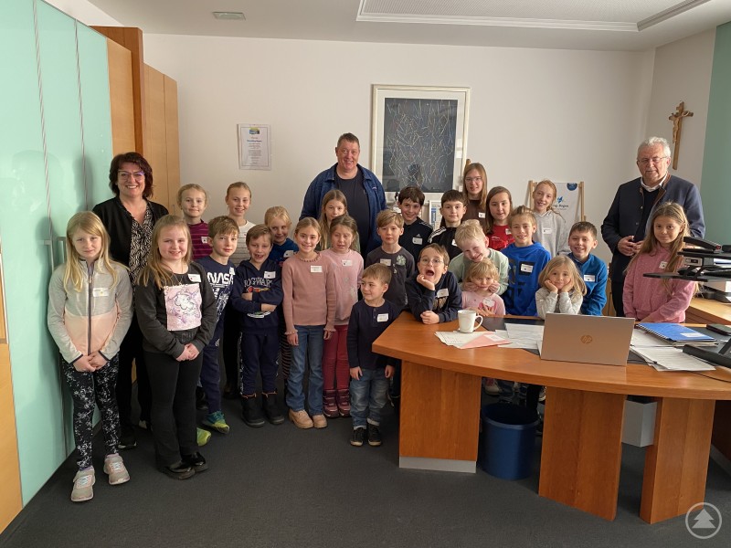 Die Kinder im Büro des Landrats gemeinsam mit der Gleichstellungsbeauftragten Michaela Kunze-Venus, dem Personalratsvorsitzenden Alexander Penzkofer und Landrat Josef Laumer Eine große Gruppe von Kindern steht gemeinsam mit drei erwachsenen Personen in einem Büro. Die Kinder lächeln in die Kamera und tragen Namensschilder. Hinter ihnen befinden sich ein Schreibtisch, Schränke und ein Kreuz an der Wand.