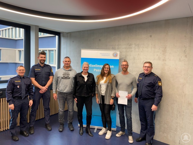 Bild v.l.n.r.: EPHK Michael Ammerl, POM Stefan Schinke, Johann Dickl, Manuela Dickl, Katja Obernhuber, Christian Gottschalk, PD Christian Dichtl Sieben Personen stehen in einer Reihe in einem modernen Raum mit großen Fenstern und Betonwand. Drei davon sind Polizeibeamte in Uniform, die übrigen vier Zivilpersonen. In der Mitte hält ein Mann ein Ehrenschreiben. Hinter der Gruppe steht ein Banner der Polizeiinspektion Passau.