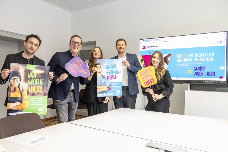 Vorstellung der digitalen Willkommensplattform Niederbayern in Landshut v.l.n.r. Alexander Zendelbach Niederbayern Forum, Dr. Reinhard Saller Geschäftsführer Niederbayern Forum, Carina Weckmüller idowapro, Felix Happe Wirtschaftsförderer der Stadt Landshut, Vanessa Becker Projektleiterin idowapro Fünf Personen stehen in einem modernen Besprechungsraum und halten bunte Plakate mit Slogans wie Work hier und Einfach ankommen in die Kamera. Im Hintergrund ist eine Präsentationsfolie zur digitalen Willkommensplattform Niederbayern zu sehen.
