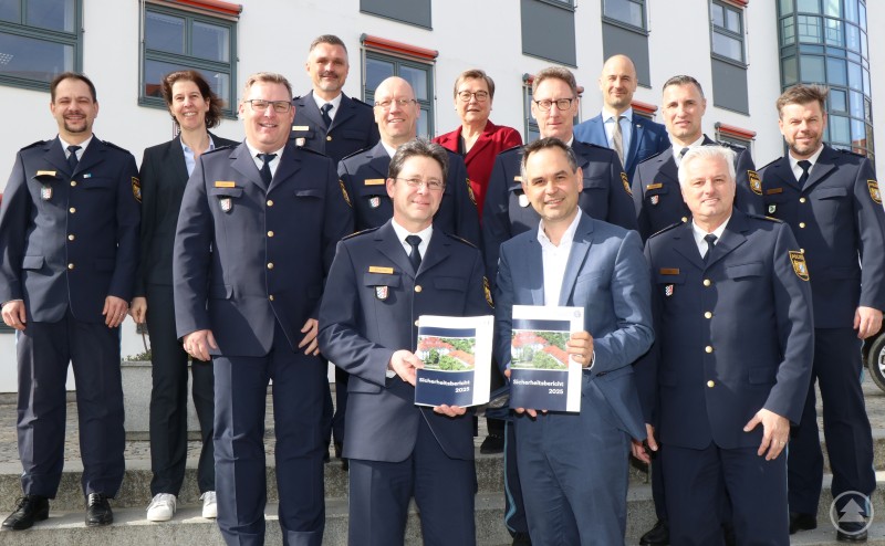 Polizeipräsident Roland Kerscher (vorne links), Polizeivizepräsident Werner Sika (vorne rechts) und Leitende Kriminaldirektorin Annette Haberl (dritte, letzte Reihe) zusammen mit Landrat Raimund Kneidinger (vorne mittig) und Leiterin der Abteilung Sicherheit und Ordnung, Karin Sedlmaier, (hinten mittig) sowie den Dienststellenleiterinnen und Dienststellenleitern der zuständigen Polizeidienststellen im Landkreis Passau bei der Vorstellung des Sicherheitsberichtes. Mehrere Führungskräfte der Polizei sowie Vertreter des Landratsamtes stehen und sitzen vor einem Gebäude für ein Gruppenfoto. Zwei Personen halten den Sicherheitsbericht 2025 in die Kamera.