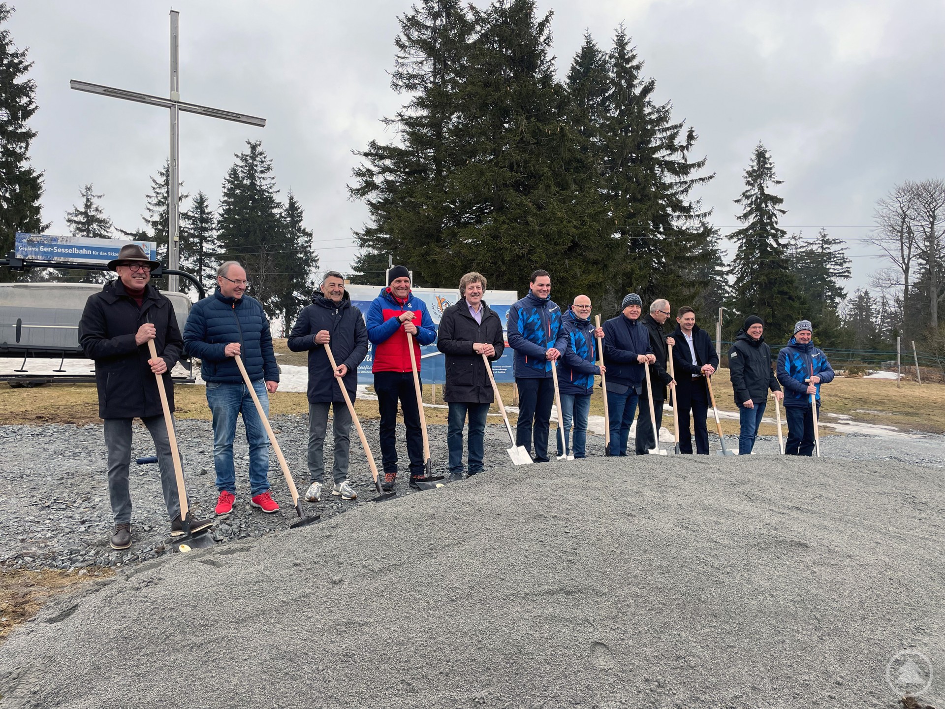 Zwölf Personen stehen nebeneinander vor einem kleinen Kieshaufen und halten Spaten in der Hand – ein klassischer Spatenstich. Die Szene spielt sich im Freien ab, bei bewölktem Himmel und kühlem Wetter. Im Hintergrund erkennt man eine Sessellift-Anlage, ein großes Metallkreuz sowie schneebedeckte und schneefreie Flächen – typisch für ein Wintersportgebiet wie das Skizentrum Mitterdorf. Einige Personen tragen auffällige blaue Funktionsjacken mit Muster, was auf eine Zugehörigkeit zum Skigebiet oder einem Verein schließen lässt. Die Stimmung ist feierlich und gemeinschaftlich, passend zum symbolischen Baubeginn eines neuen Projekts.