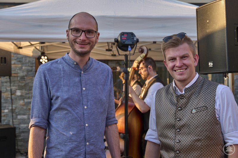 Zwei Männer lächeln in die Kamera, im Hintergrund eine Musikgruppe beim Auftritt im Festzelt.