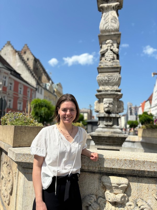Preisträgerin Anna Dachs vor dem Stadtbrunnen in der Straubinger Altstadt. Junge Frau mit weißer Bluse steht lächelnd vor einem historischen Brunnen mit kunstvoll gemeißelten Steinfiguren, im Hintergrund Altstadtgebäude und blauer Himmel mit wenigen Wolken.