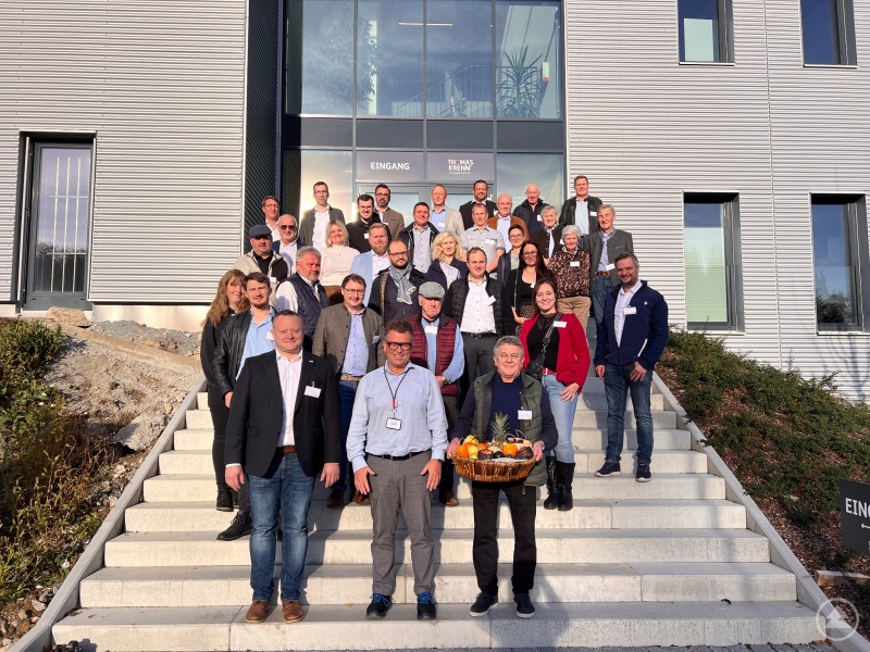 Der MU-Kreisvorsitzende FRG, Stefan Behringer (vorne links), bedankt sich bei Thomas Frei (vorne Mitte), dem COO der Thomas-Krenn.AG in Freyung, für die Führung und die interessanten Einblicke. Eine große Gruppe von Männern und Frauen steht bei Sonnenschein auf der Treppe vor dem modernen Gebäude der Thomas-Krenn.AG. In der vorderen Reihe halten zwei Männer einen Präsentkorb mit Obst, alle lächeln in die Kamera.
