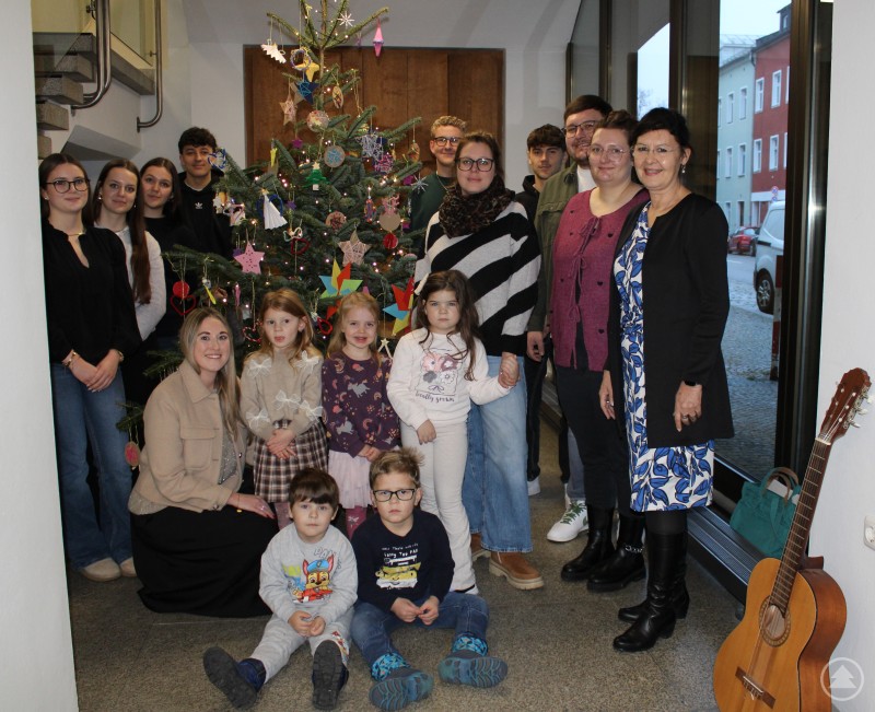 Eva-Maria Kelch (Vorsitzende der Geschäftsführung der Agentur für Arbeit Passau), Lydia Winkler (Erzieherin), Dominik Wiesmann (Leiter Kinderhaus), Tamara Schreiner (Erzieherin), Laura Fuller (stellvertretende Leiterin des Kinderhauses) mit den Kindergartenkindern, umringt von den sechs Auszubildenden der Agentur für Arbeit Passau (Sophia Unfried, Régi Boglárka, Marie-Johanna Jasker, Marko Hrskanovic-Matic, Maximilian Schlig, Jannik Berndorfer) Vor einem festlich geschmückten Weihnachtsbaum stehen und sitzen Kindergartenkinder, Auszubildende sowie Mitarbeitende der Agentur für Arbeit Passau und des Caritas Kinderhauses St. Michael. Im Vordergrund sind bunte Weihnachtsdekorationen und eine Gitarre zu sehen. Die Gruppe lächelt für das gemeinsame Foto.