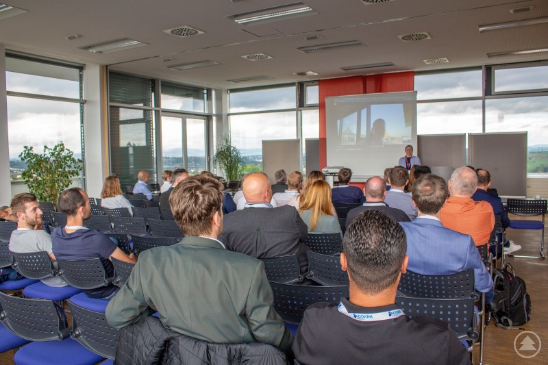 Präsentation beim Digital Tag 2024: Expertinnen und Experten diskutieren live und virtuell über Digitalisierungstrends. Menschen sitzen in einem Konferenzraum beim Digital Tag und verfolgen eine Präsentation mit Videostream auf Leinwand.