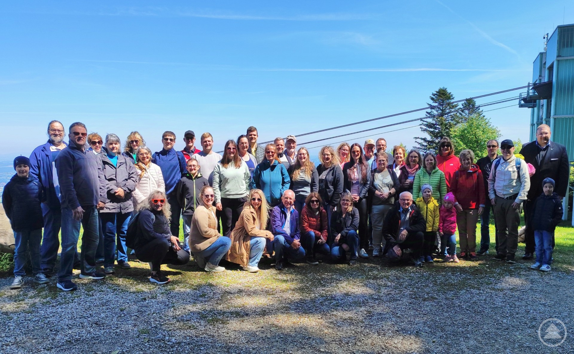 Eine große Reisegruppe des Hotsvereins 1892 Freyung steht vor der Bergstation der Grünbergseilbahn auf einem sonnigen Plateau. Im Hintergrund sind die Seilbahn und ein bewaldeter Berghang zu sehen.