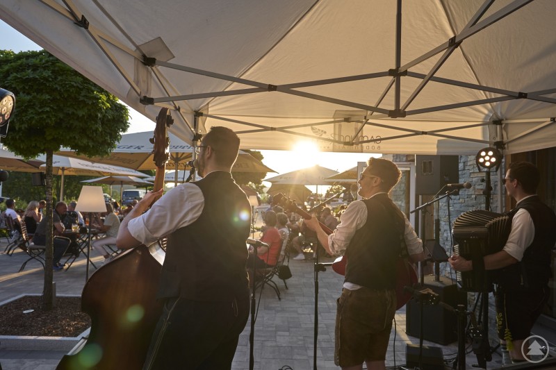 Drei Musiker spielen auf einer kleinen Bühne im Freien, dahinter sitzen Gäste bei untergehender Sonne.
