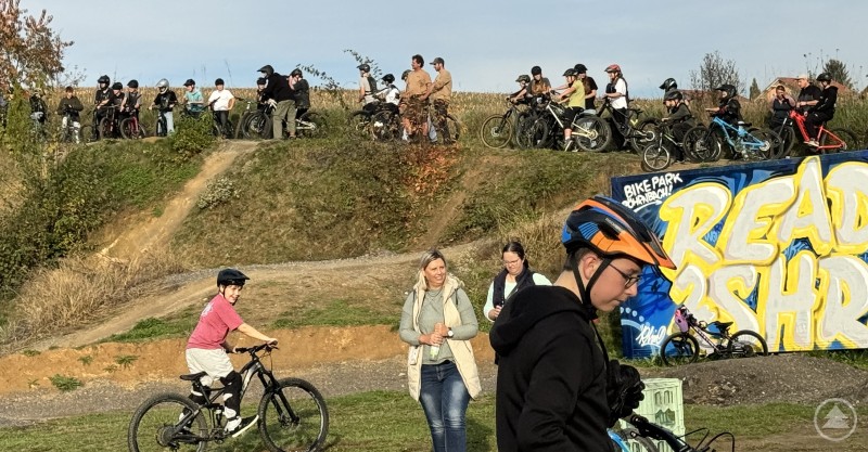 Beim „Woid Summa goes Bike – Dreck & Gaudi“ in Röhrnbach steht Spaß und Bewegung für Jugendliche im Vordergrund. Junge Radfahrerinnen und Radfahrer mit Helmen versammeln sich auf einem Hügel im Bikepark, im Vordergrund ein Junge auf dem Fahrrad sowie Zuschauerinnen – im Hintergrund bunte Graffitiwand mit „READY 2 SHRED“.
