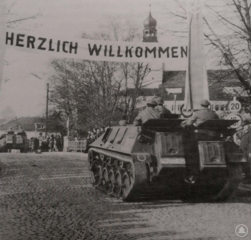 Schwarz-weiß Foto mit historischen Militärfahrzeugen, die unter einem Banner mit der Aufschrift 