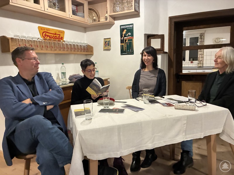 Bei der Buchpräsentation im Paul-Friedl-Haus stellten Herausgeber, Verlegerin und Übersetzerin den neuen Band „Mein Weg zu unseren Deutschen“ vor. Vier Personen sitzen an einem Tisch im Paul-Friedl-Haus, unterhalten sich und lesen aus einem Buch. Auf dem Tisch liegen mehrere Bücher und Gläser.