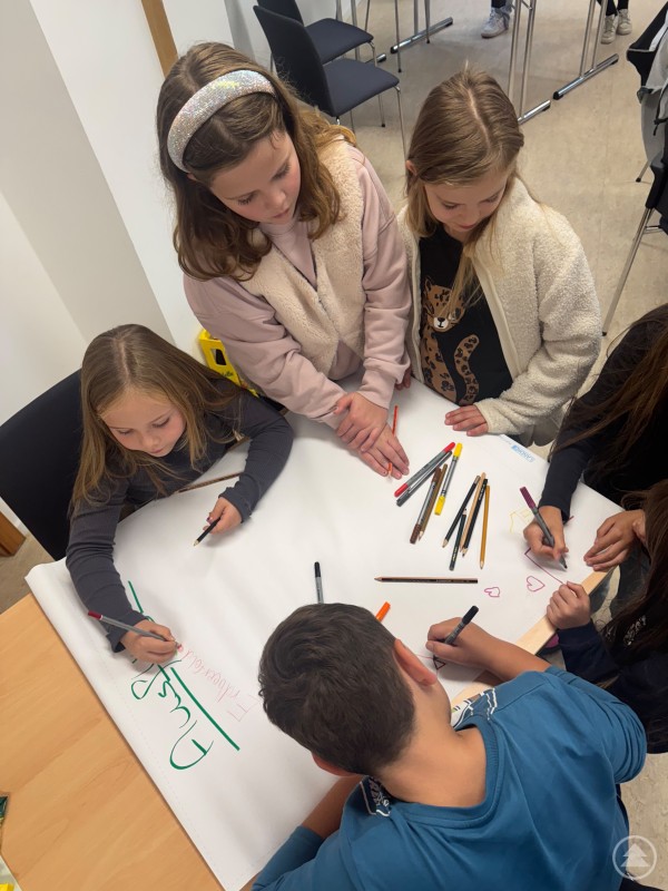 Mit viel Liebe gestalteten die Kinder ein Plakat mit ihren Wünschen. Mehrere Kinder stehen an einem Tisch und gestalten mit bunten Stiften ein großes Plakat.