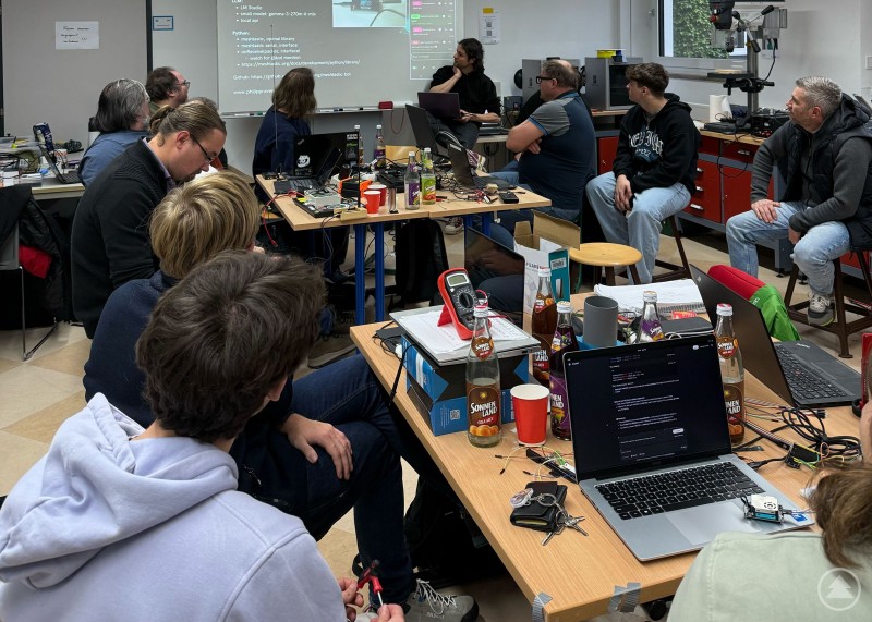 Mehrere Teilnehmer sitzen in einer Werkstatt an Tischen voller Laptops, Messgeräte und Elektronikbauteile. Auf einer Leinwand läuft eine technische Präsentation zu Python und LM Studio.