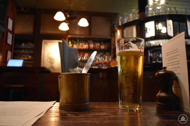 Ein frisch eingeschenktes Bierglas steht auf einem Holztisch in einem gemütlichen Wirtshaus. Im Hintergrund sind eine Bar, warme Lampen und zahlreiche Flaschen zu sehen. Neben dem Bier stehen Besteck in einem Behälter sowie eine Speisekarte.
