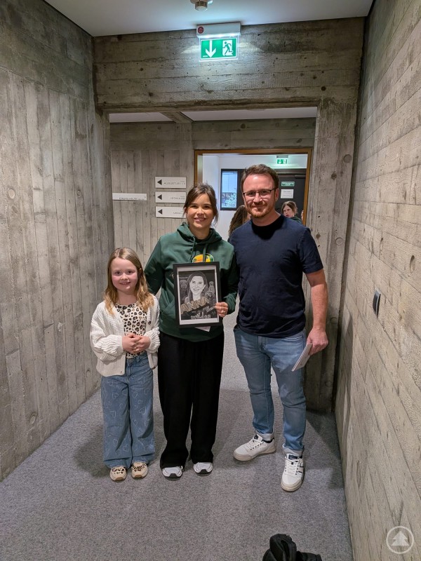 Annika Preil freut sich über das detailreiche Portrait von Florian Eibl Annika Preil hält gemeinsam mit einem Künstler und einem Kind ein gezeichnetes Portrait in einem Flur