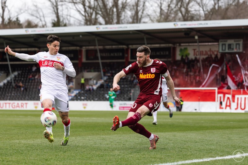 Zwei Fu&szlig;ballspieler k&auml;mpfen auf dem Spielfeld um den Ball, im Hintergrund sind Trib&uuml;ne und Fans zu sehen