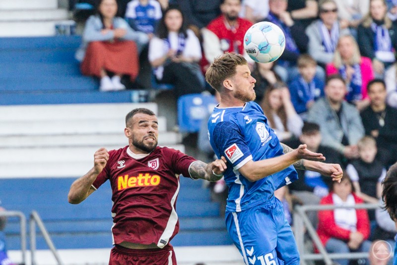 Auf dem Bild ist eine dynamische Fußballszene zu sehen: Zwei Spieler kämpfen im Kopfballduell um den Ball. Der Spieler im blauen Trikot scheint den Ball mit dem Kopf zu treffen, während der Spieler im weinroten Trikot mit „Netto“-Werbung auf der Brust in engem Kontakt mit ihm steht. Im Hintergrund sind viele Zuschauer auf den Tribünen zu erkennen, die das Spiel aufmerksam verfolgen. Das Bild vermittelt Spannung und Intensität des Spiels.