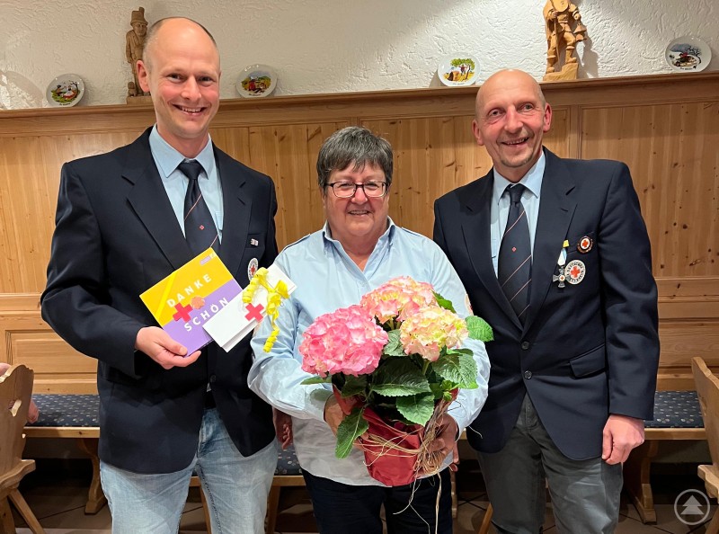 Eine Frau mit Blumen und zwei Männer in BRK-Uniform posieren lächelnd vor einer Holzvertäfelung; einer hält eine Dankeskarte.