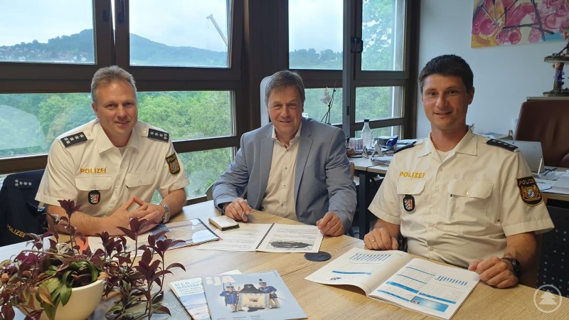 v.l.n.r. EPHK Kern, Bürgermeister Alexander Mayer, PHK Franz Weinzierl Zwei Polizeibeamte in weißer Uniform und ein Bürgermeister im Anzug sitzen an einem Besprechungstisch mit Unterlagen und Broschüren, im Hintergrund große Fenster mit Blick ins Grüne.