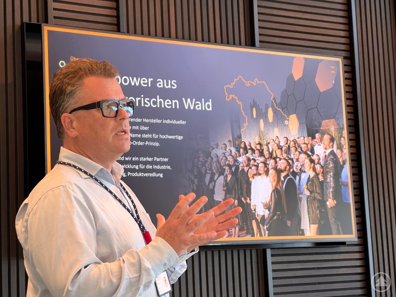 Ein Mann mit Brille hält einen Vortrag vor einem großen Bildschirm. Auf dem Bildschirm sind eine Karte von Bayern, viele Personen und der Slogan „Serverpower aus dem Bayerischen Wald“ zu sehen.