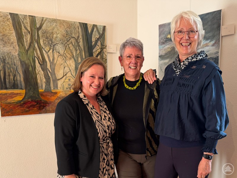 Drei Frauen stehen l&auml;chelnd vor mehreren gro&szlig;formatigen Gem&auml;lden in einer Ausstellung und pr&auml;sentieren die Werke der K&uuml;nstlerin.