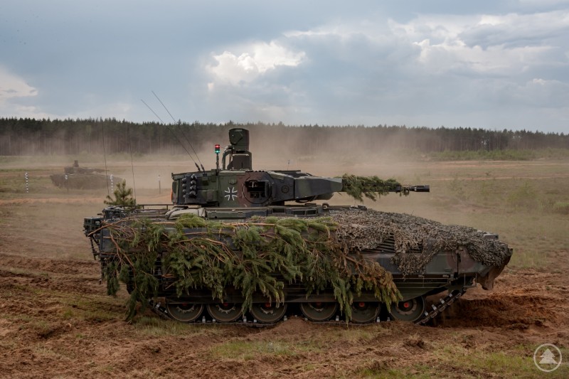 Zwei getarnte Schützenpanzer Puma der Bundeswehr im Manöver auf staubigem Feld, mit Wald im Hintergrund.