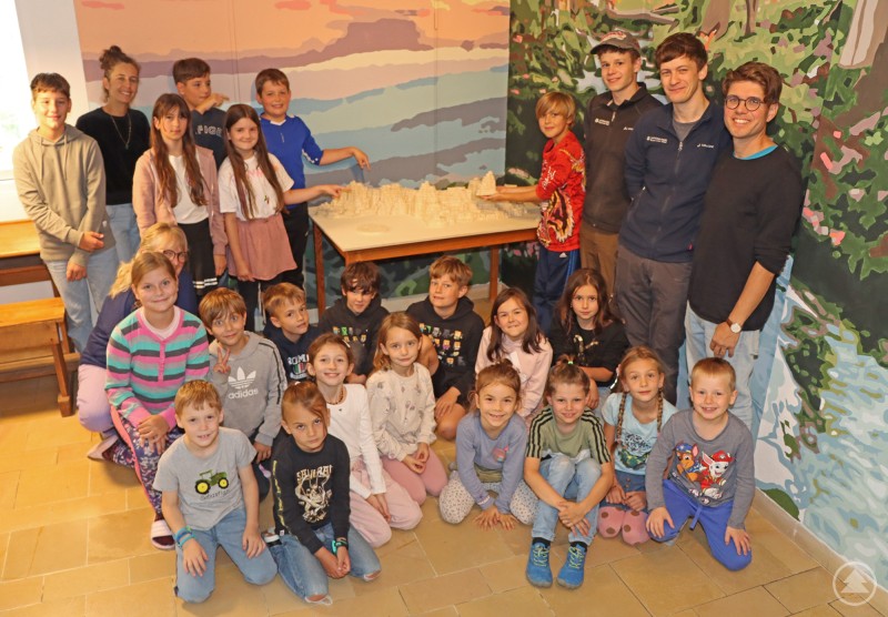 Die Kinder und Lehrer der Montessori-Schule Freyung mit Jan Kiesling (hinten, 2. v. r.) und Jeremias Großmann vom Nationalpark Bayerischer Wald. Eine Gruppe von Kindern und Jugendlichen posiert gemeinsam mit mehreren Erwachsenen vor einem Modell des Nationalparks, das auf einem Tisch in einem bunt bemalten Raum steht. Die Kinder sitzen oder knien vorne, die älteren stehen im Hintergrund.