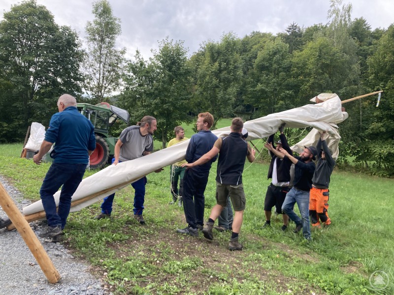 Das Regionalbudget 2025 brachte erneut frischen Wind in die Region. Ein Beispielprojekt ist in Ringelai: Dort errichtete der Orts-Caritasverein mit tatkräftiger ehrenamtlicher Unterstützung ein großes Tipi, das rund 20 Kindern und Jugendlichen als neuer Begegnungsort dient. Mehrere Männer arbeiten gemeinsam im Freien daran, ein großes, weißes Tipi-Zelt aufzustellen. Im Hintergrund sind Bäume, eine Wiese und ein Traktor zu sehen.