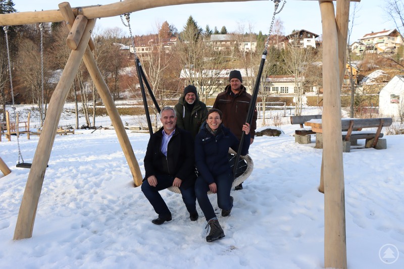 Freuen sich über den neu gestalteten Wanderpark: Bürgermeister Michael Herzog (v.l.), Robert Kürzinger, Geschäftsführer der Ferienregion Nationalpark Bayerischer Wald, Nationalparkleiterin Ursula Schuster und Nationalparkmitarbeiter Martin Weiderer. Vier Erwachsene stehen und sitzen gemeinsam auf einer großen Schaukel im verschneiten Wanderpark von Bayerisch Eisenstein. Im Hintergrund sind weitere Spielgeräte, Schnee und Häuser zu sehen. Die Stimmung ist freundlich und winterlich.