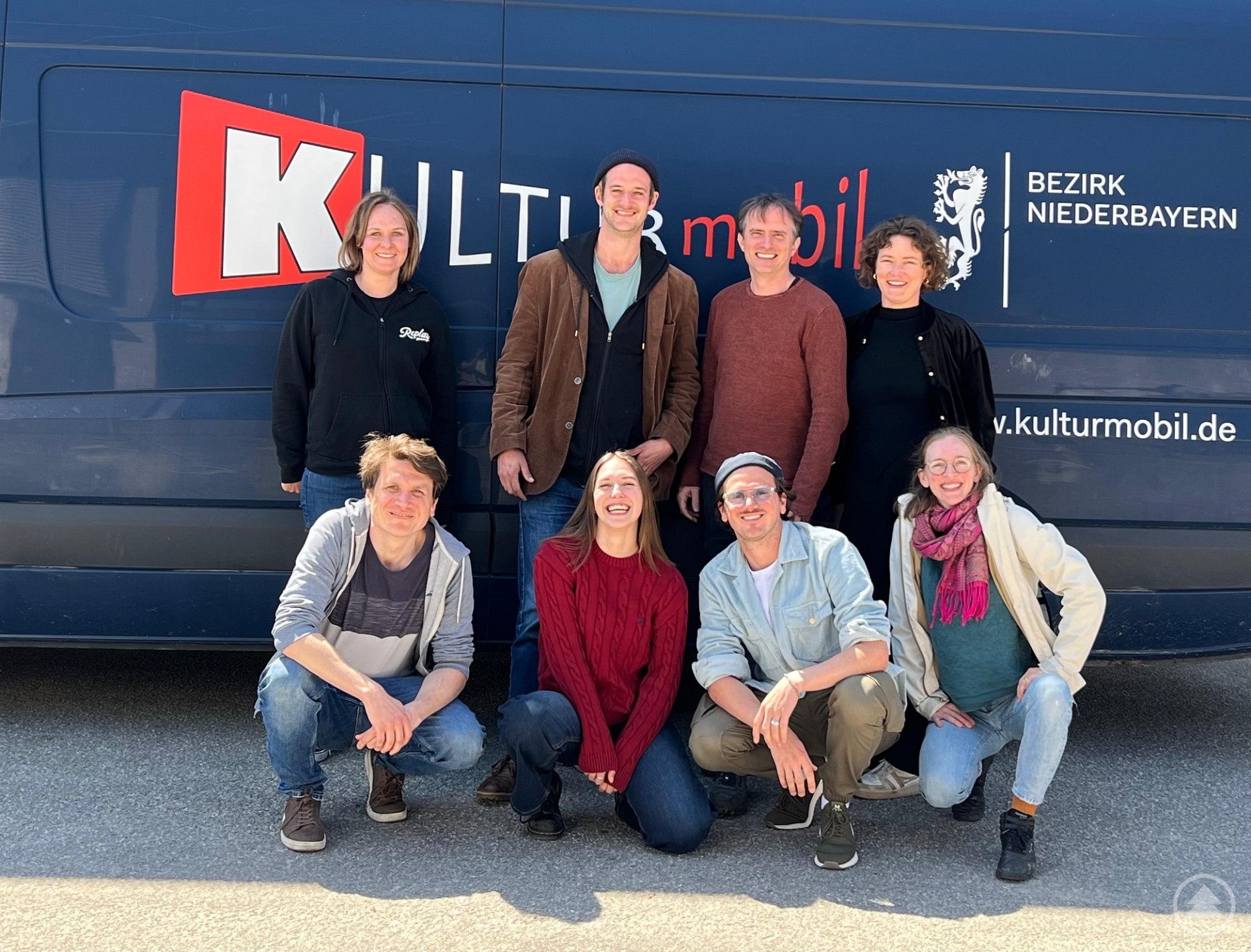 Das KULTURmobil-Team posiert vor einem dunkelblauen KULTURmobil-Bus mit dem Logo des Bezirks Niederbayern. Acht Teammitglieder stehen und sitzen lächelnd vor dem Bus.